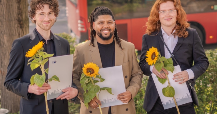 Drei glückliche junge Männer mit ihrem Abschluss und Sonnenblumen in den Händen