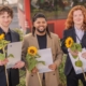 Drei glückliche junge Männer mit ihrem Abschluss und Sonnenblumen in den Händen
