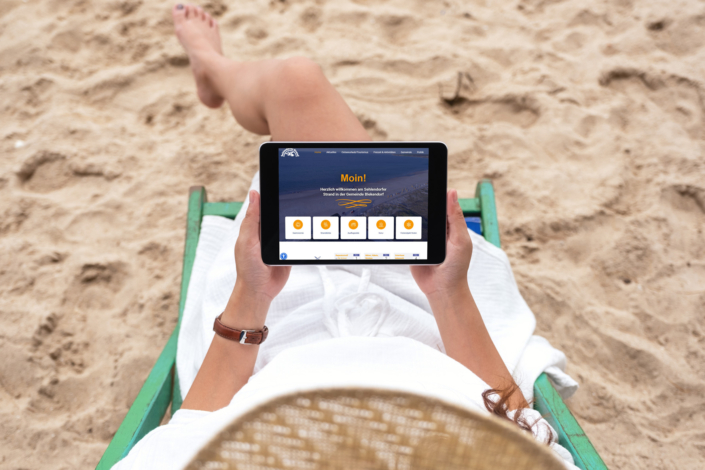 Eine Frau mit Strohhut sitzt am Strand und betrachtet die neue Website der Gemeinde Blekendorf auf einem Tablet